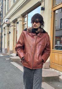 Homme portant une veste en cuir marron, une écharpe rayée, des lunettes de soleil et des boucles d'oreilles, debout sur un trottoir en ville près de portes de boutique en bois.