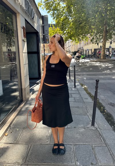 Top negro sin mangas, falda midi negra, zapatos planos negros. Sosteniendo un bolso de mano tejido naranja. Entorno al aire libre con árboles y elementos de la calle de fondo.