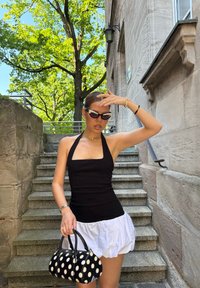 Top negro de halter, falda blanca con volantes y bolso negro con lunares. Las gafas de sol y las joyas añaden detalles distintivos. Fondo de escalera de piedra.