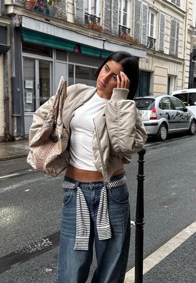 Chaqueta bomber beige, camiseta blanca, jeans anchos azules con un cinturón a rayas y un bolso tote de estampado de serpiente texturizado. Entorno urbano.