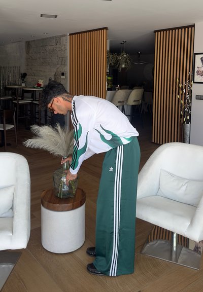 Persona con chándal verde de Adidas con rayas blancas interactúa con un jarrón de cristal que contiene plantas secas, al lado de una mesa circular de madera y hormigón.