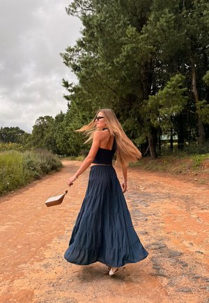 Mujer de cabello largo con gafas de sol, blusa negra y falda azul fluida caminando por un sendero de tierra rodeado de árboles.