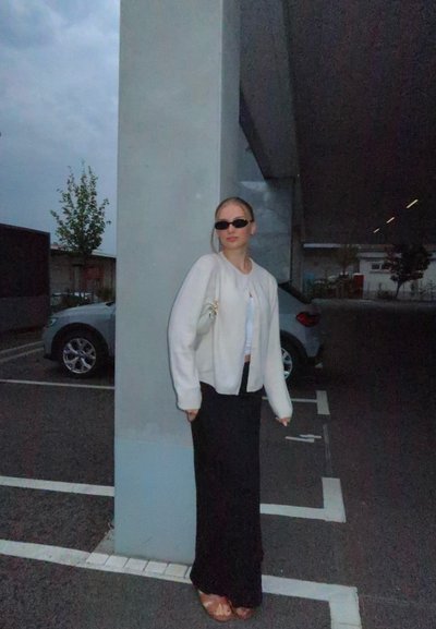 Chaqueta bomber blanca, falda maxi negra y sandalias marrones. De pie contra un pilar de concreto, llevando gafas de sol, bajo un cielo nublado.