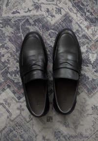 Črne usnjene loafers čevlje s ploskim, okroglem prstom in dekorativnim šivanjem. Gladka tekstura z minimalnimi dodatki, postavljeni na vzorčasto preprogo.
