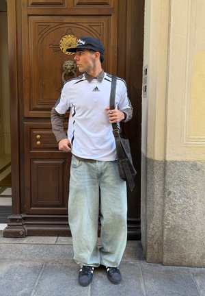 Joven con una camiseta blanca de Adidas, camiseta de manga larga a rayas debajo, vaqueros holgados, gorra negra y bolso de hombro, de pie junto a una puerta de madera.