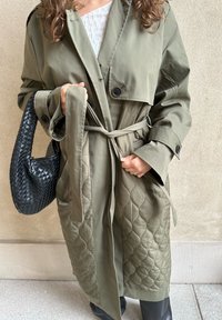 Ελαιοπράσινο παλτό trench με μαξιλαράκια, κουμπιά και ζώνη, συνδυασμένο με ένα υφασμάτινο μαύρο τσάντα. Λείο ύφασμα, μεσαίου μήκους, μεγάλες μανίκες.
