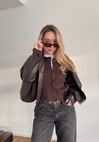 Jeune femme aux longs cheveux blonds ajustant des lunettes de soleil noires, portant une veste en cuir marron, un pull en tricot, un jean foncé et portant un sac tressé.