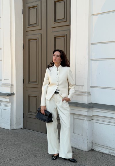 Mujer con un traje color marfil sosteniendo un bolso de mano negro, de pie con una mano en el bolsillo frente a una gran puerta marrón y una pared blanca.