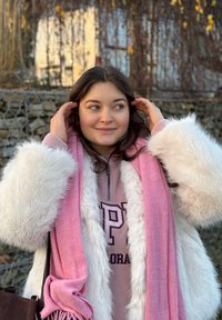 Cappotto di pelliccia bianca, sciarpa rosa, felpa grigia con logo. Capelli lunghi e castani, espressione dolce, in piedi all'aperto vicino a un muro di pietra.