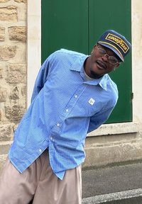 Ριγέ μπλε πουκάμισο με κουμπιά, μπεζ παντελόνι και ναυτικό καπέλο με το κεντημένο "GOODYEAR". Τοποθετημένο μπροστά σε έναν πέτρινο τοίχο και μια πράσινη πόρτα.