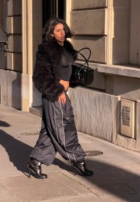 Femme marchant sur le trottoir en ville, portant un crop top noir, une veste duveteuse, un pantalon large, des bottes à talons hauts, et tenant un sac à main noir.