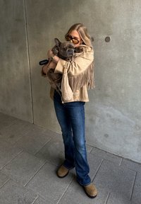 Bežna jakna s resicami in dolgimi rokavi, temne kavbojke in rjave klompe. Drži sivo francosko buldogko. Ozadje prikazuje sivo betonsko steno.