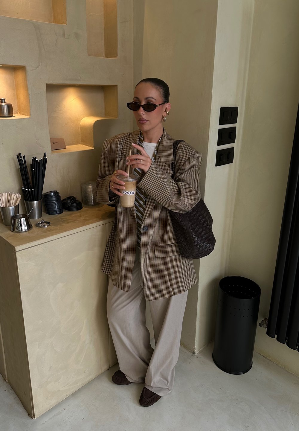 Blazer oversized în carouri maro, cămașă albă cu dungi verticale, pantaloni bej deschis, ținând o cafea rece. Geantă texturată maro, elemente negre pe perete.
