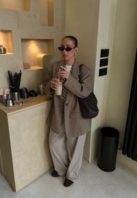 Blazer oversized în carouri maro, cămașă albă cu dungi verticale, pantaloni bej deschis, ținând o cafea rece. Geantă texturată maro, elemente negre pe perete.