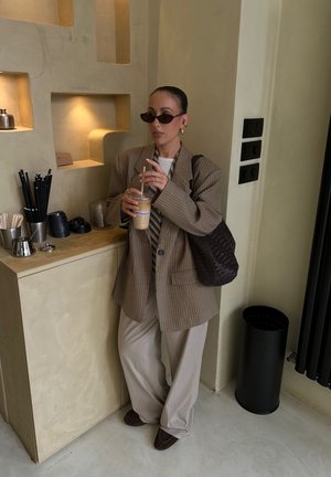 Braunes karierter übergroßer Blazer, weißes Hemd mit vertikalen Streifen, hellbeige Hose, hält eine Eiskaffee. Strukturiert braune Tasche, schwarze Wandelemente.
