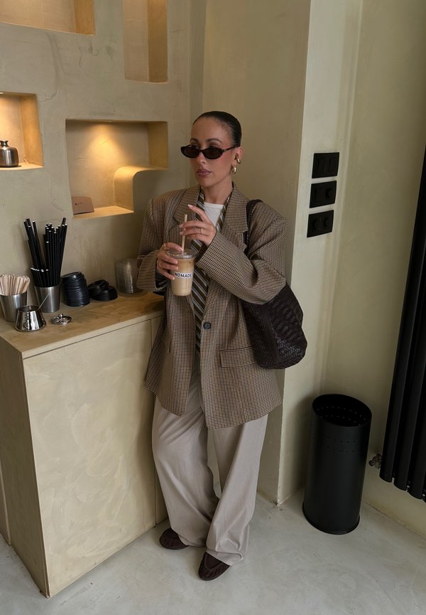 Blazer oversize à carreaux marron, chemise blanche à rayures verticales, pantalon beige clair, tenant un café glacé. Sac en cuir texturé marron, éléments muraux noirs.