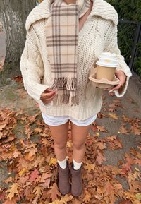 Suéter de punto grueso crema con cuello chal, bufanda a cuadros beige, pantalones cortos blancos y botas marrones. Sosteniendo una taza de café en un portavasos de cartón.