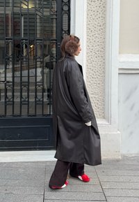 Femme portant un long manteau noir, des cache-oreilles marron, un pantalon large foncé et des chaussures rouges et blanches, debout sur un trottoir près d'une grille métallique et d'un mur.