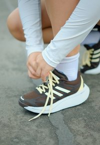 Persoon in witte lange mouwen die gele veters straktrekt op bruin en witte hardloopschoenen op een asfaltoppervlak.