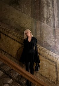 Jeune femme aux cheveux blonds, en robe noire et gants, debout sur un escalier en pierre orné, devant un mur fresqué ancien et détaillé, tenant un sac noir.
