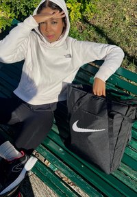 Geantă tote Nike neagră cu logo-ul swoosh alb, înconjurată de un hanorac gri deschis, colanți negri și adidași negri cu accente roșii.