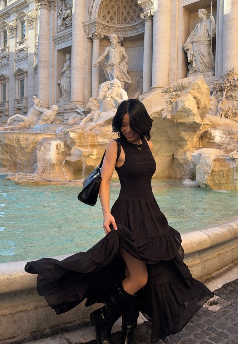 Vrouw in een vloeiende zwarte jurk en laarzen draait rond bij de Trevifontein, met klassieke standbeelden en turquoise water op de achtergrond.