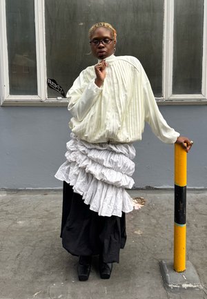 Blouse blanche plissée, jupe blanche à volants superposée sur une longue jupe noire, assortie à des chaussures plateforme noires. Poteau jaune et noir en arrière-plan.