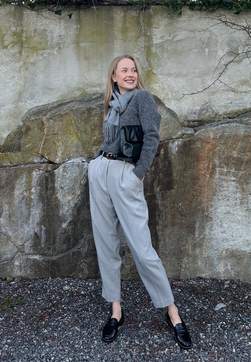 Femme en pull gris, écharpe, pantalon clair et mocassins noirs, debout souriante contre un mur de pierre recouvert de mousse, les mains dans les poches.