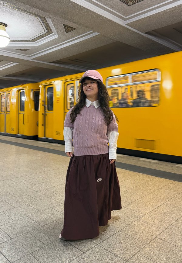 Jeune femme en pull rose et casquette se tient sur un quai de métro avec un train jaune qui passe derrière elle.