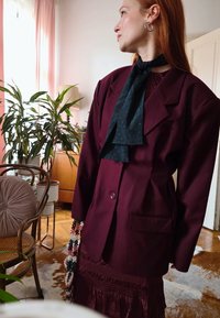 A woman with red hair is wearing a dark burgundy jacket and dress, a black scarf, and hoop earrings. She is standing in a room that features plants and wooden furniture.