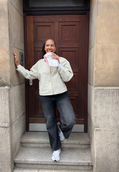 Chaqueta de color claro con cremallera sobre una camiseta oscura, jeans oscuros y zapatillas blancas. Sosteniendo un alimento en una bolsa de papel, de pie junto a una puerta de madera.