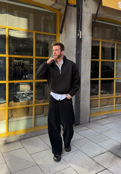 Hombre con suéter negro y pantalones negros anchos camina por la acera afuera de un café con ventanas de marco amarillo y asientos al aire libre.