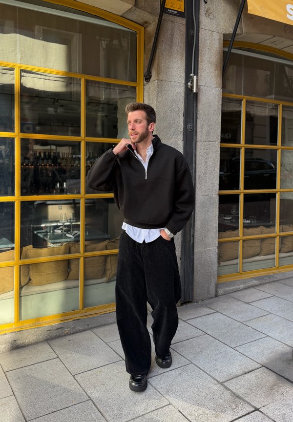 Un homme portant un pull noir et un pantalon large noir marche sur le trottoir devant un café avec des fenêtres encadrées de jaune et une terrasse extérieure.