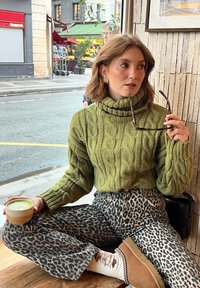 Groene kabelgebreide turtleneck trui, wijde broek met luipaardprint, beige instappers, houdt een groene drank in een beker vast, zit binnen.