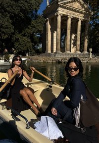 To kvinner sitter i en liten hvit båt på vannet. Den ene har på seg en svart kjole, den andre et denimantrekk. Begge har solbriller. I nærheten er en historisk steinstruktur.