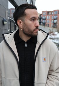 Homme aux cheveux noirs et barbe portant plusieurs boucles d'oreilles en or, un pull noir zippé et une veste beige Carhartt, debout à l'extérieur près de bâtiments.