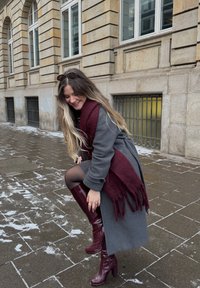 Jeune femme en manteau gris et écharpe bordeaux ajustant ses bottes bordeaux montantes jusqu'aux genoux sur un trottoir enneigé d'une ville, près d'un bâtiment en pierre avec de grandes fenêtres.