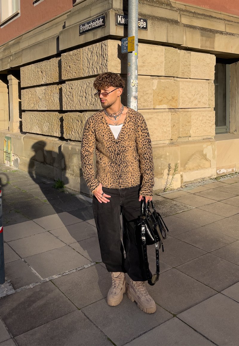 Cardigan com padrão de leopardo, camiseta branca por baixo, calças largas pretas, botas chunky bege e uma bolsa preta; em pé num passeio perto de um edifício.
