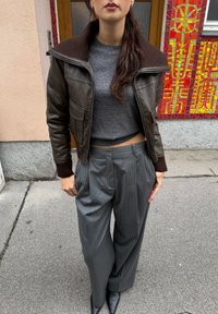 Hnědá kožená bomber bunda s ribbed límcem, šedý zkrácený svetr a šedé široké kalhoty s proužky. Černé boty s špičatou špičkou.