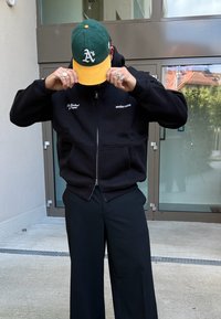 Persoană ajustând o șapcă de baseball verde și galben, purtând un hanorac negru cu fermoar cu text alb și pantaloni negri largi, stând în fața unor uși de sticlă.
