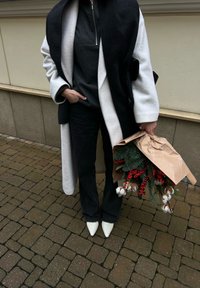 Personne portant un manteau noir et blanc et des chaussures blanches pointues tenant un bouquet enveloppé dans du papier brun avec du feuillage, des baies rouges et des tiges de coton.