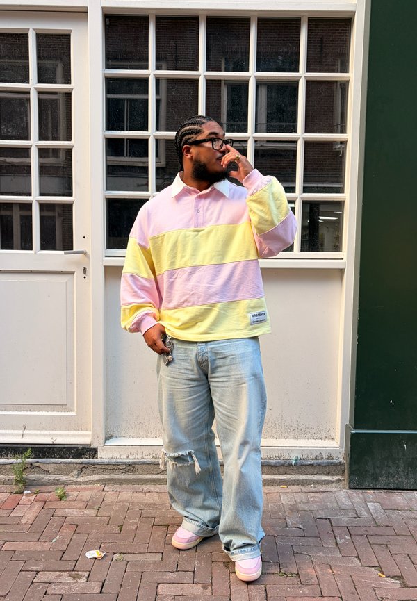 Man with braided hair and glasses wearing a pink and yellow striped shirt, ripped jeans, and pink shoes standing on brick pavement.