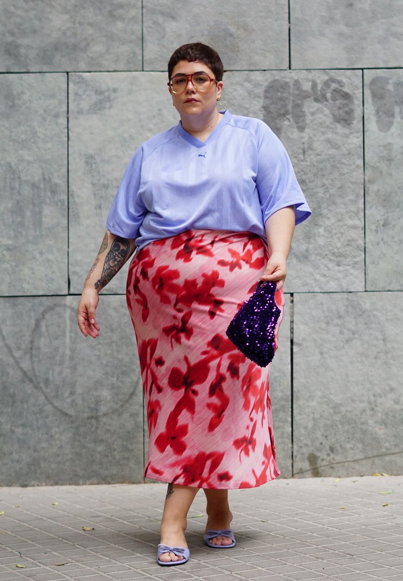 Personne aux cheveux courts portant des lunettes, une chemise violet clair, une jupe fleurie rouge, des sandales violettes et tenant un petit sac violet à paillettes.