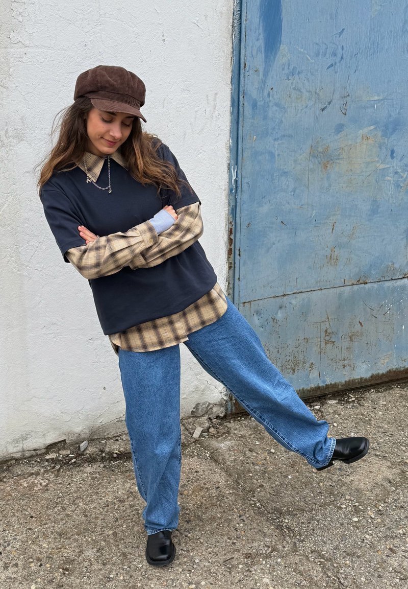 Giovane donna con berretto marrone, camicia a quadri stratificata e top nero, jeans blu e scarpe nere, in piedi su una gamba accanto a un muro bianco e una porta blu.