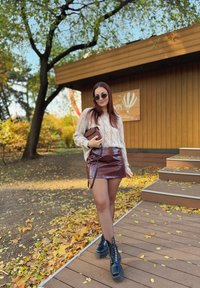 Kabelstrickcreme Pullover, bordeauxroter, glänzender Minirock, schwarze Schnürstiefel und runde Sonnenbrille. Herbstliches Laub in einer Parklandschaft.