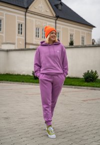 Henkilö kävelee ulkona purppuranvärisessä urheiluasuissa, oranssissa pipoissa ja keltaisissa lenkkareissa, hymyillen kädet taskuissa.