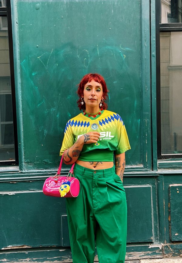 Persona con cabello rojo que lleva una camiseta amarilla y verde de Brasil, pantalones verdes y sostiene un bolso rosa con un llavero amarillo, de pie contra una pared verde.