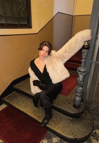 Νεαρή γυναίκα με μαύρο φόρεμα και μπεζ γούνινο παλτό που κάθεται σε πέτρινα σκαλοπάτια, κρατώντας ένα μαύρο τσαντάκι, με το χέρι της να ακουμπάει στη διακοσμητική κουπαστή της σκάλας.