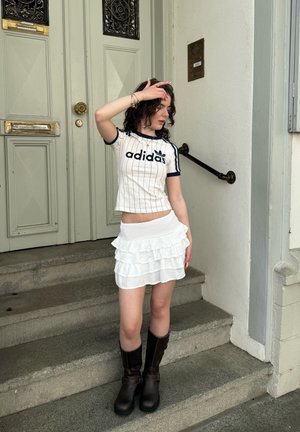 Balti Adidas trumpas viršus su tamsiai mėlynomis juostelėmis ir logotipu, derinamas su sluoksniuotu baltu raukšlėtu mini sijonu ir rudos spalvos aukštakulniais batais iki kelių.