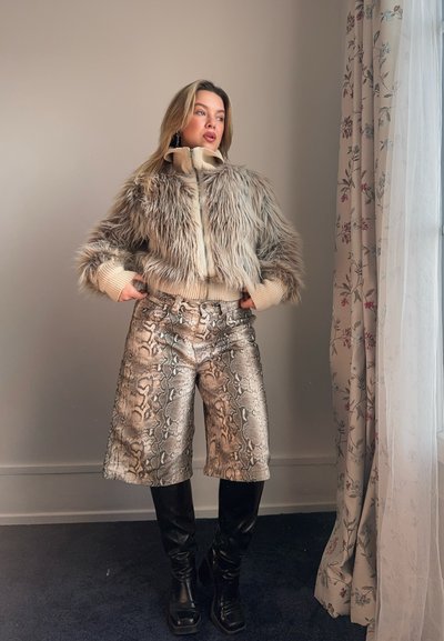 Chaqueta corta de piel sintética en beige y gris con puños acanalados, combinada con pantalones cortos metálicos de estampado de serpiente y botas negras hasta la rodilla.